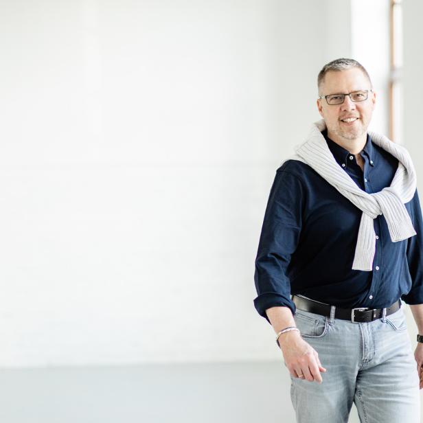 Mann mit Brille und Pullover über den Schultern steht in einem hellen, modernen Raum.