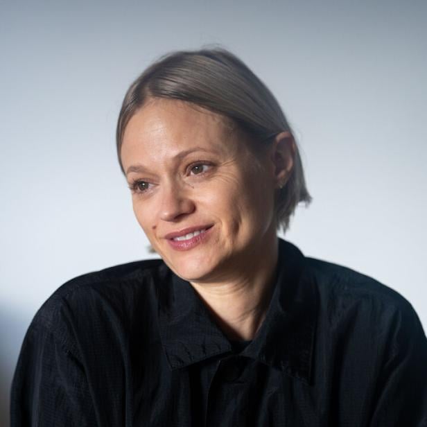 Frau mit kurzem, blondem Haar trägt eine schwarze Jacke und lächelt leicht, während sie die Arme verschränkt.