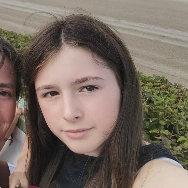 Ein Mädchen mit langen braunen Haaren steht draußen und schaut direkt in die Kamera.