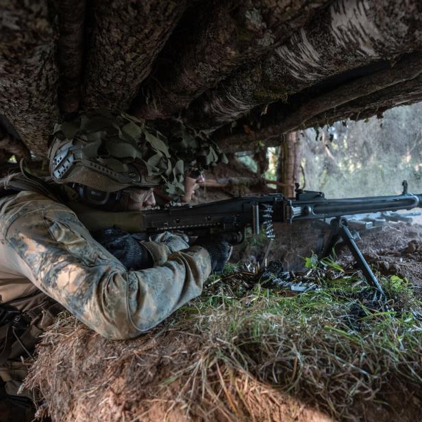 Ein Soldat in Tarnkleidung zielt mit einem Maschinengewehr aus einer getarnten Deckung im Wald.