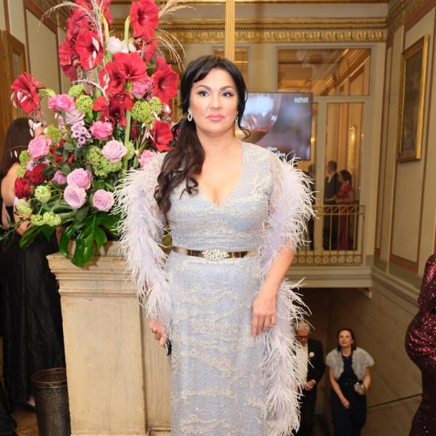 Eine Frau in einem eleganten silbernen Abendkleid mit Federn steht vor einem großen Blumenarrangement in einem prunkvollen Saal.