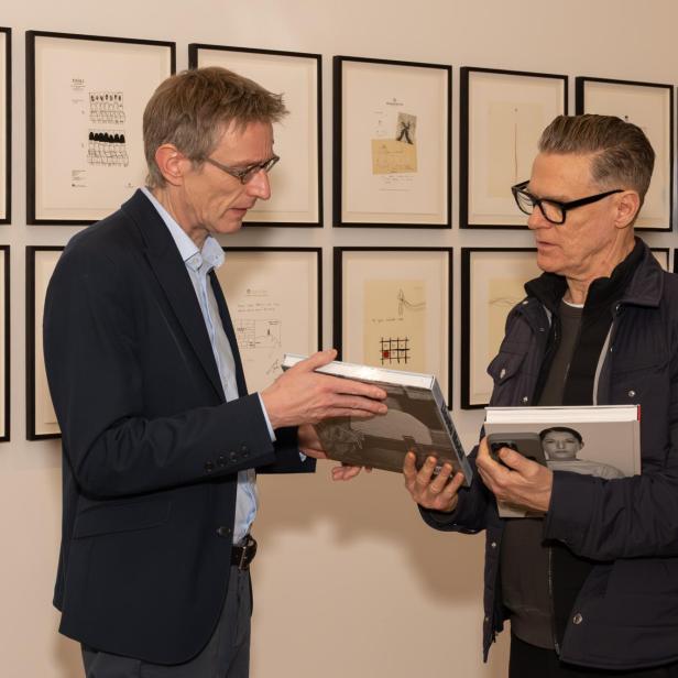 Zwei Männer unterhalten sich und halten Bücher in den Händen, im Hintergrund hängen gerahmte Zeichnungen an der Wand.