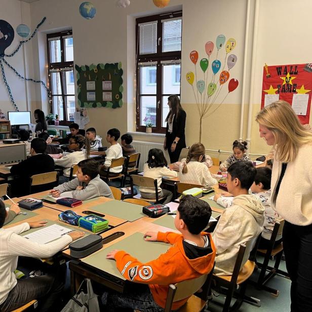 Viele Kinder sitzen an Tischen in einem Klassenraum, während zwei Erwachsene sie betreuen und unterrichten.