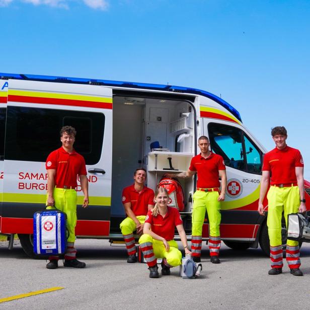 Fünf Rettungskräfte in rot-gelber Kleidung stehen und sitzen vor einem geöffneten Rettungswagen auf einer Landstraße.