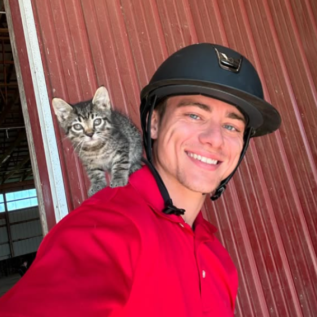Mann in rotem T-Shirt steht mit einer Katze auf seiner Schulter und einem schwarzen Reithelm auf dem Kopf vor einer Wand