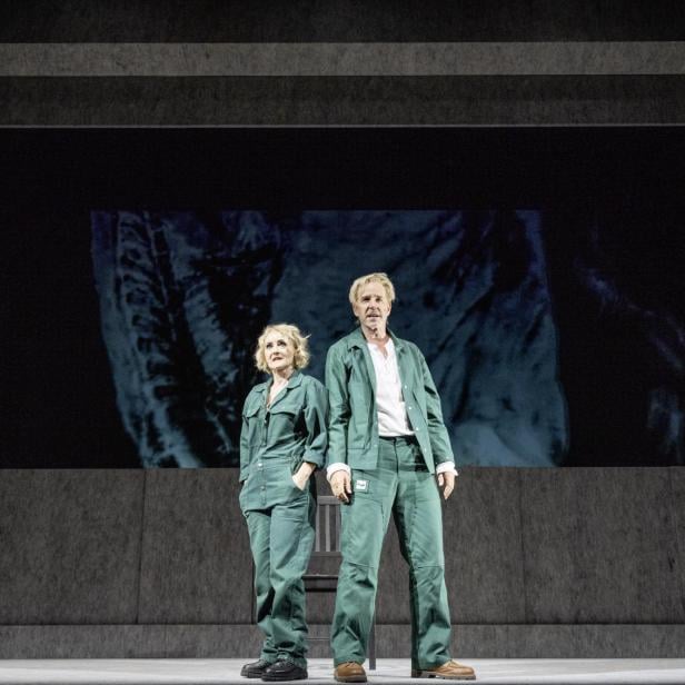 Zwei Personen in grüner Arbeitskleidung stehen auf einer Bühne vor einer großen, dunklen Leinwand.