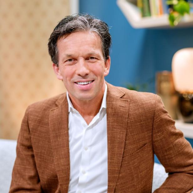 Ein Mann mit braunem Sakko und weißem Hemd sitzt lächelnd auf einem Sofa in einem modern eingerichteten Raum.