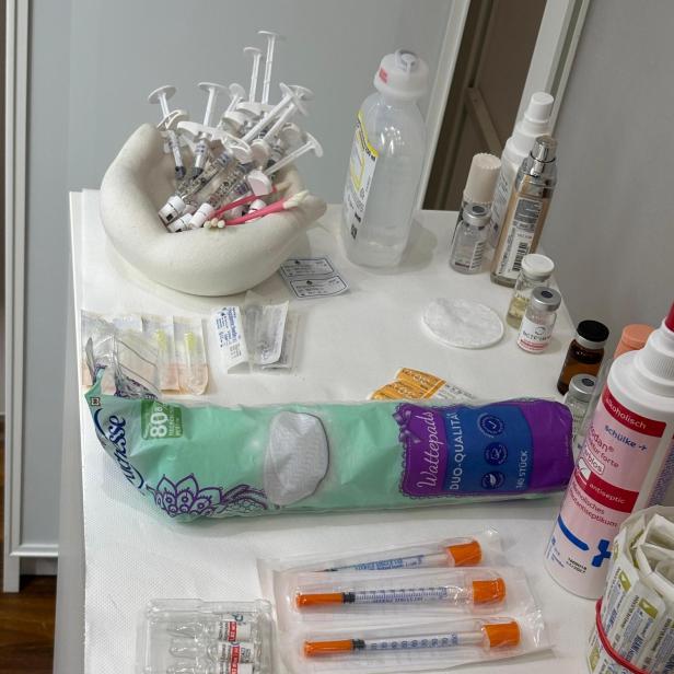 Mehrere Spritzen, Ampullen, Desinfektionsmittel, Wattepads und medizinische Utensilien liegen auf einer weißen Ablage.