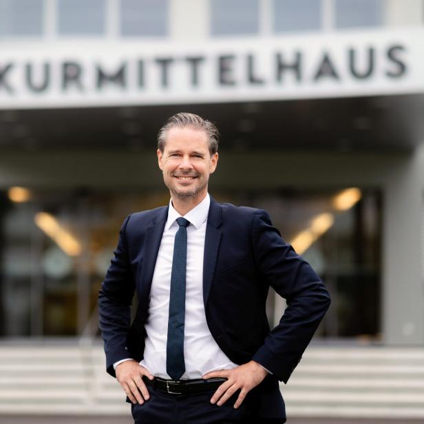 Ein Mann im Anzug steht lächelnd mit Händen in der Hüfte vor einem Gebäude mit der Aufschrift „Kurmittelhaus“.