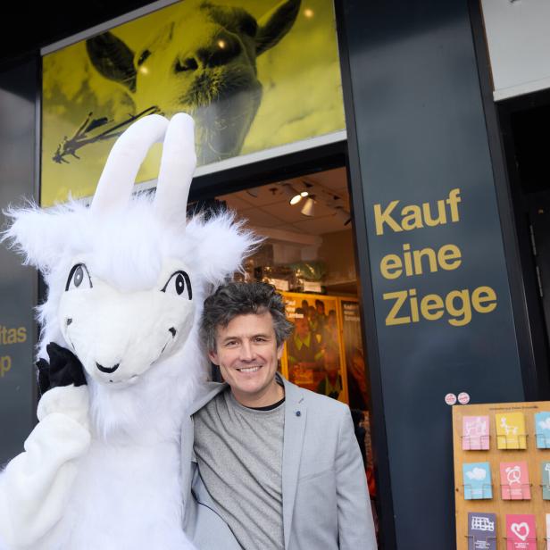Ein Mann lächelt neben einem Ziegen-Maskottchen vor einem Laden mit der Aufschrift "Kauf eine Ziege".