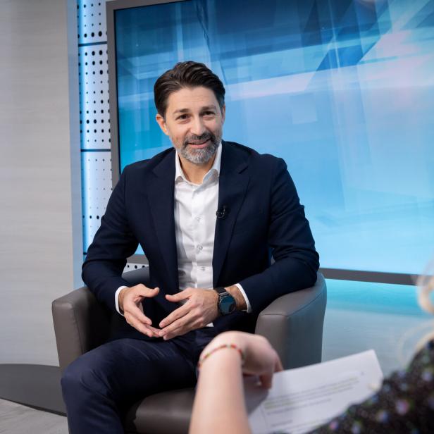 Ein Mann im Anzug sitzt in einem modernen Studio und spricht mit einer Frau, die ihm Fragen stellt.