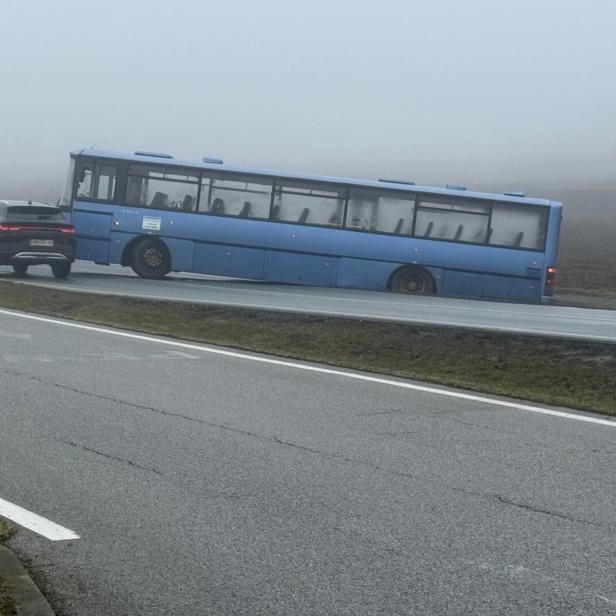 Ein blauer Bus steckt schräg im Straßengraben, während Autos auf einer nebligen Straße vorbeifahren.
