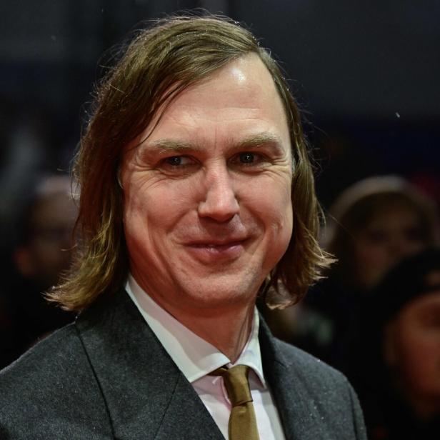 Ein Mann mit mittellangem braunem Haar trägt Anzug und Krawatte und lächelt in einer Menschenmenge.