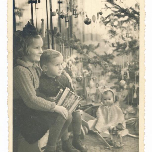 Drei Kinder sitzen neben einem geschmückten Weihnachtsbaum, eines hält ein Akkordeon, im Hintergrund liegt Spielzeug.