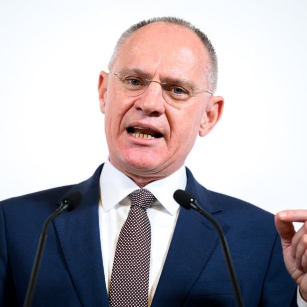 Ein Mann im Anzug spricht gestikulierend vor zwei Mikrofonen vor hellem Hintergrund.