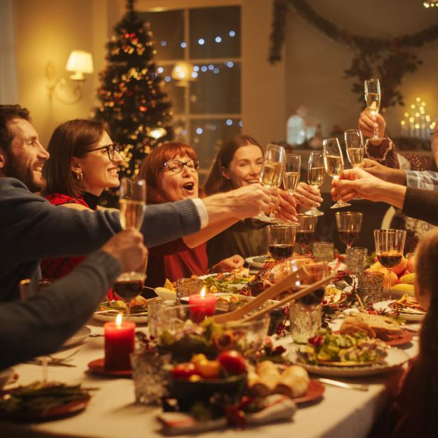 Mehrere Menschen stoßen an einem festlich gedeckten Tisch mit Sektgläsern an, im Hintergrund steht ein geschmückter Weihnachtsbaum.