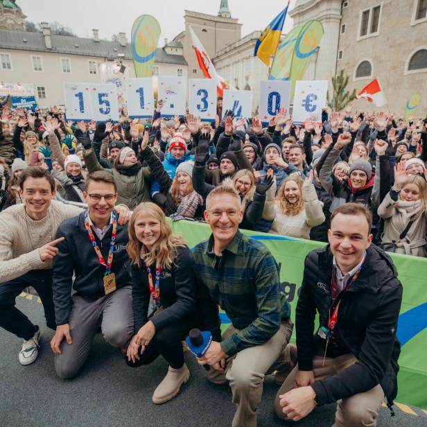Fünf Personen knien vor einer jubelnden Menschenmenge, die Schilder mit der Spendensumme 151.340 € hochhält.
