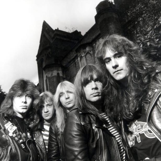 Fünf Männer mit langen Haaren und Lederjacken stehen vor einem alten, dunklen Gebäude.