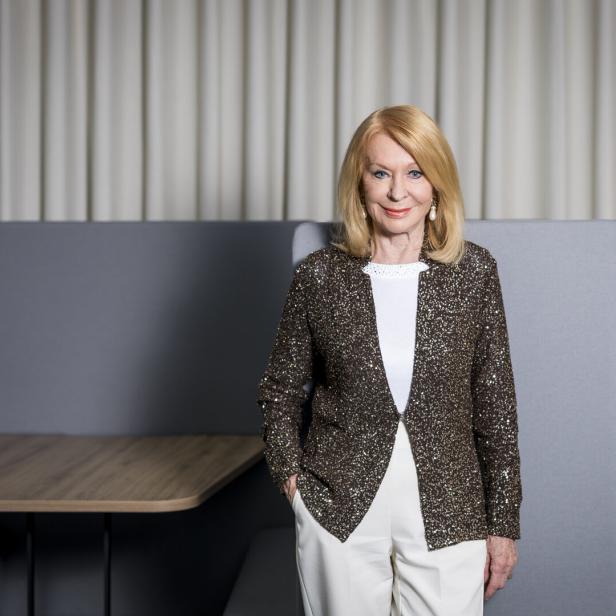 Eine Frau mit blondem Haar trägt eine glitzernde Jacke und weiße Hose, steht vor grauen Sitzmöbeln und hellem Vorhang.