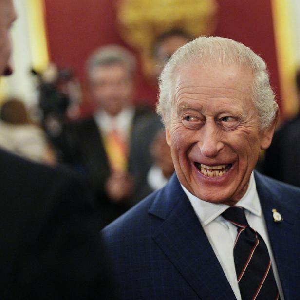 König Charles III. lacht herzlich in einem Gespräch, im Hintergrund sind weitere Personen zu sehen.