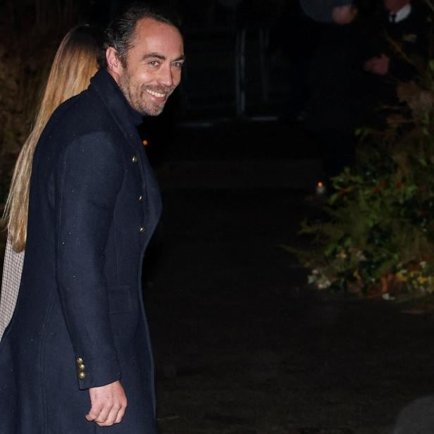 Ein Mann mit dunklem Mantel lächelt und steht im Freien neben einer Frau mit langen, blonden Haaren.