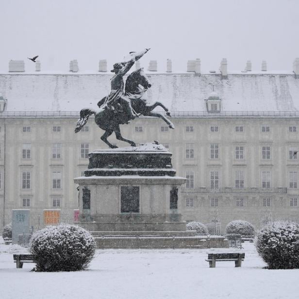 Reiterstatue auf einem verschneiten Platz mit Büschen und Bänken vor einem großen historischen Gebäude.