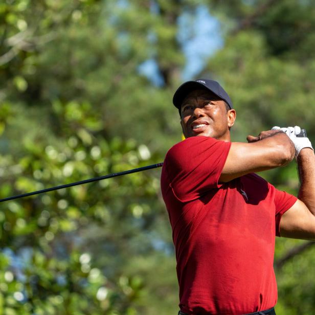 Ein Mann mit rotem Shirt und schwarzer Kappe schwingt einen Golfschläger auf einem grünen Golfplatz.