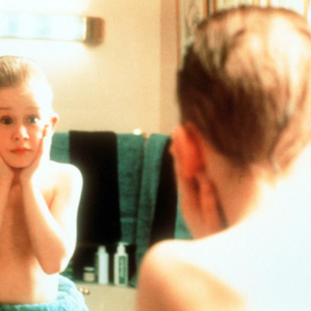 Ein Junge steht mit erschrockenem Gesichtsausdruck und Händen an den Wangen vor einem Badezimmerspiegel.