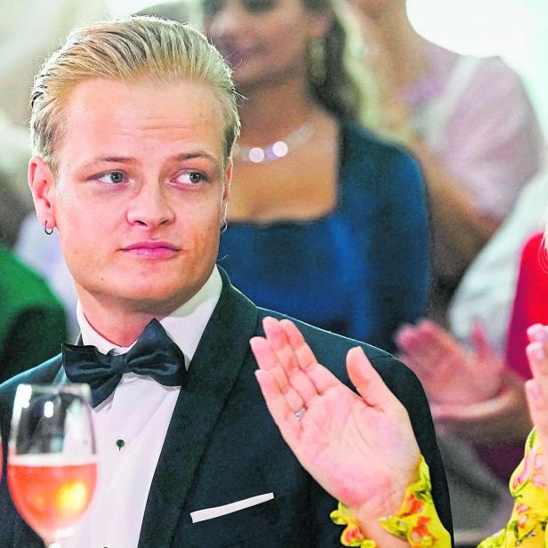 Ein junger Mann im Smoking sitzt neben einer Frau mit blondem Haar und gelbem Kleid, beide wirken festlich und aufmerksam.