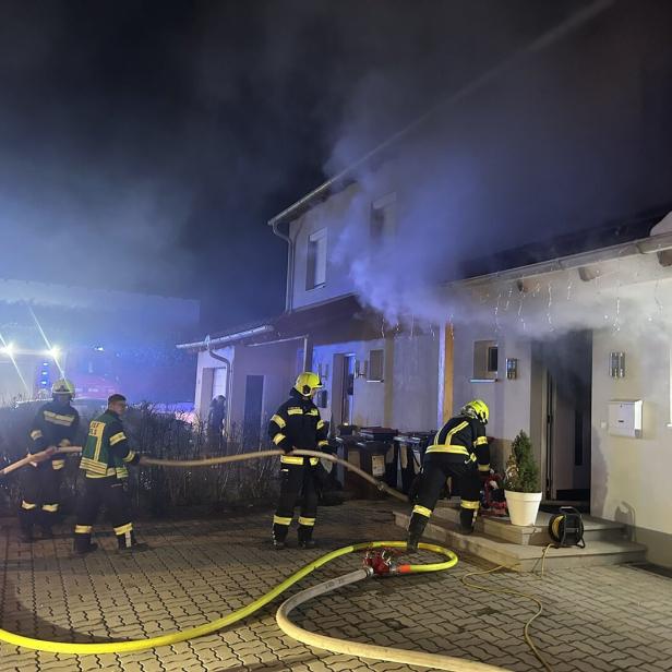 Mehrere Feuerwehrleute bekämpfen nachts einen Brand in einem Wohnhaus, aus dem dichter Rauch dringt.