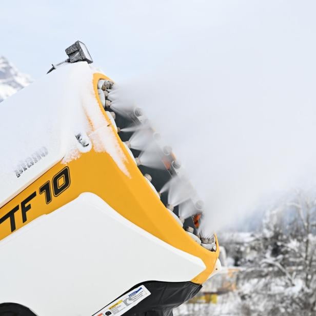 Eine gelbe Schneekanone bläst Schnee in eine winterliche Landschaft mit verschneiten Bäumen und Bergen im Hintergrund.