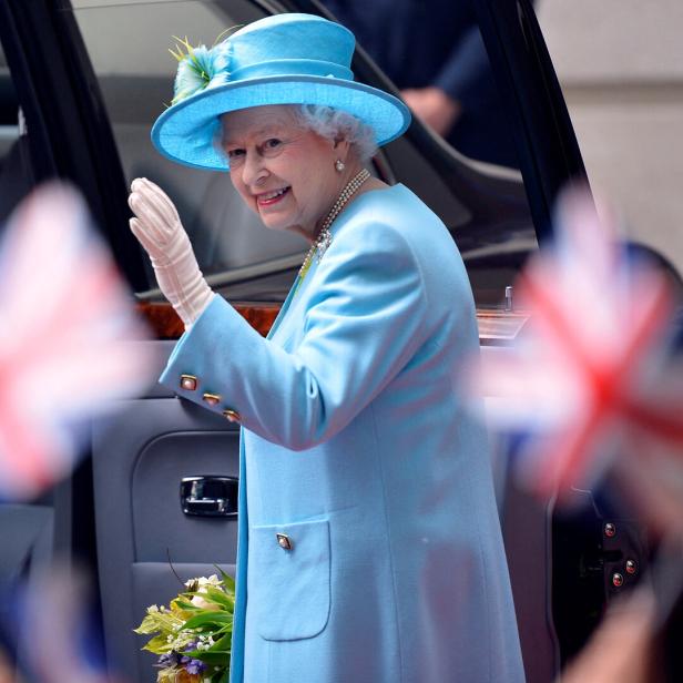 Königin Elisabeth II. in hellblauem Kostüm und Hut winkt aus einem Auto.