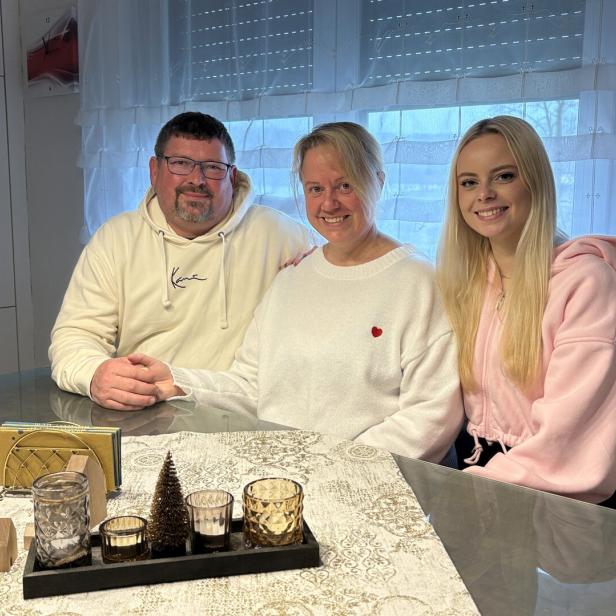 Drei Personen sitzen lächelnd an einem Tisch mit Kerzen und Dekoration, im Hintergrund ein Fenster mit Gardinen.