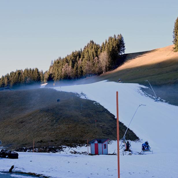 Eine schmale Skipiste mit Kunstschnee verläuft durch eine grüne Wiese, umgeben von Nadelbäumen und Schneekanonen.