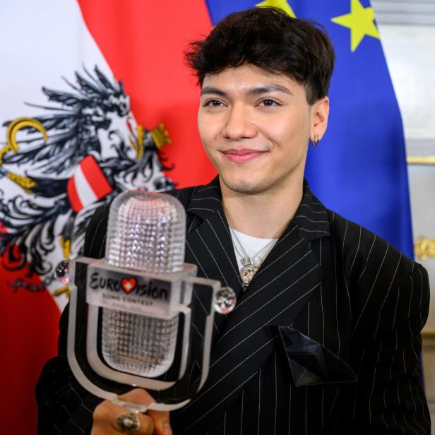 Ein junger Mann hält eine Eurovision-Trophäe vor österreichischer und EU-Flagge und lächelt.
