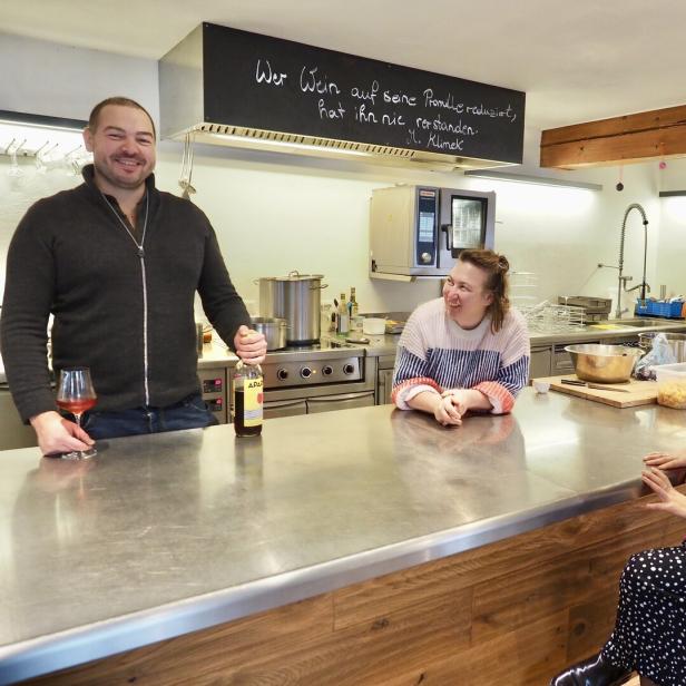 Drei Personen stehen und sitzen lächelnd an einer Küchentheke mit Getränken.