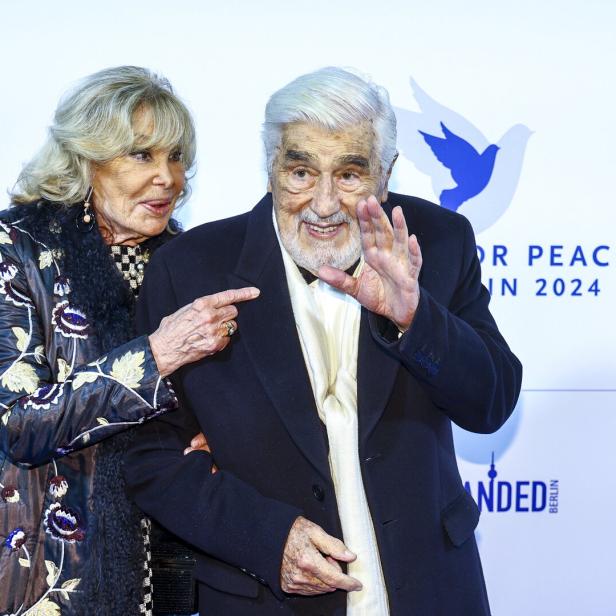 Ein älteres Paar steht lächelnd nebeneinander, während der Mann winkt und die Frau ihn am Arm hält.