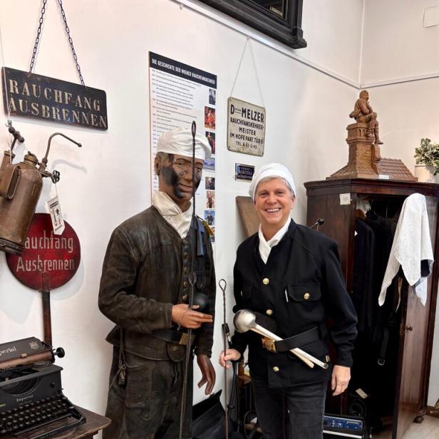 Eine Person in traditioneller Schornsteinfeger-Uniform steht lächelnd neben einer Schornsteinfeger-Figur mit Werkzeugen.