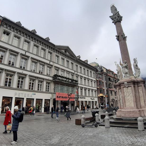 Eine belebte Fußgängerzone mit historischen Gebäuden, einer hohen Säule mit Statuen und mehreren Passanten bei bewölktem Himmel.