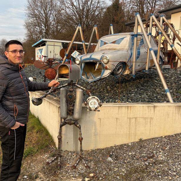 Ein Mann steht neben einer skurrilen Metallskulptur, im Hintergrund ein Auto mit Spinnenbeinen aus Metall.