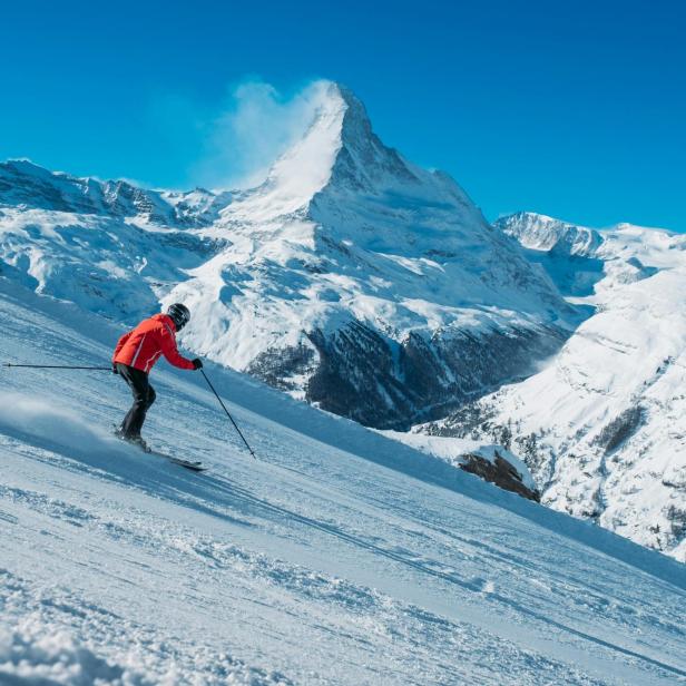 Eine Person fährt auf Skiern eine verschneite Piste hinab, im Hintergrund ragt das Matterhorn in den blauen Himmel.