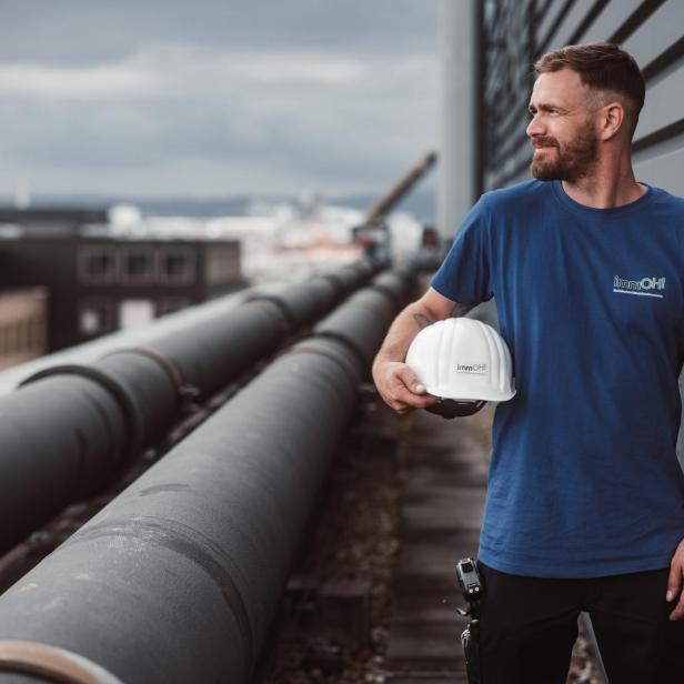 Mitarbeiter mit immOH!-Shirt hält Schutzhelm auf einem Gebäudedach neben großen Rohrleitungen.