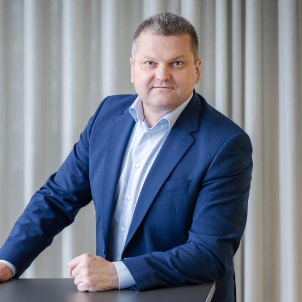 Ein Mann im blauen Anzug steht an einem Tisch vor einem hellen Vorhang und blickt ernst in die Kamera.