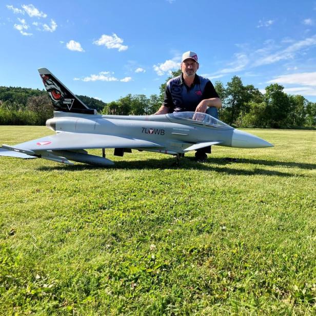 Ein Mann kniet auf einer Wiese hinter einem großen Modellflugzeug, umgeben von Bäumen und blauem Himmel.