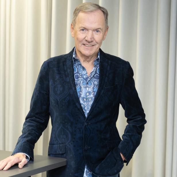 Ein lächelnder Mann mit blauem Hemd und Sakko steht vor einem hellen Vorhang.