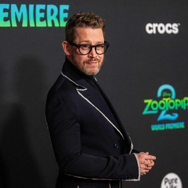 Ein Mann mit Brille und Bart steht vor einer Wand mit dem Zootopia-2-Logo und schaut zur Seite.