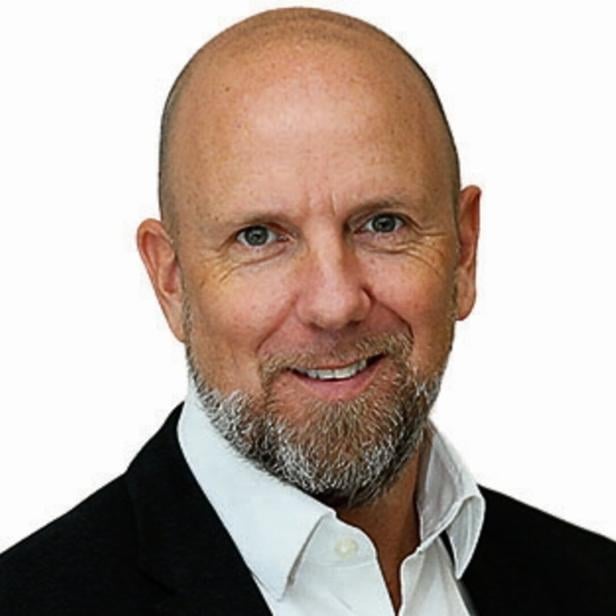 Ein Mann mit Glatze, grauem Bart und Anzug lächelt freundlich vor weißem Hintergrund.