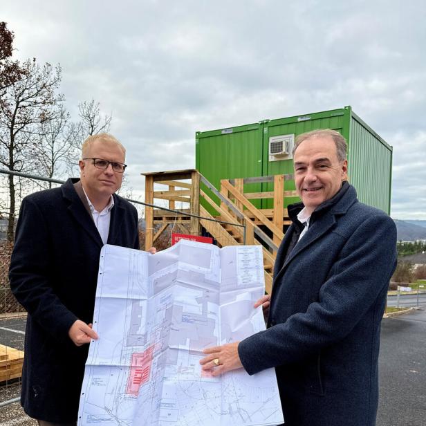 Zwei Männer im Mantel halten gemeinsam einen großen Bauplan vor einem grünen Container mit Holztreppe.