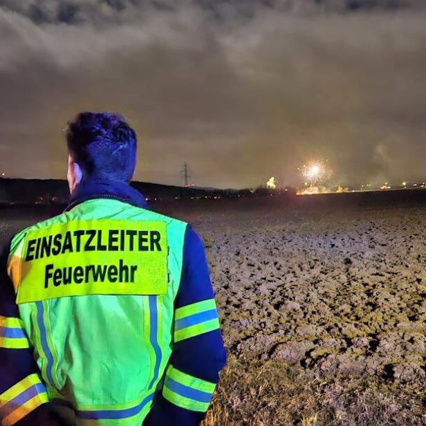 Eine Person in Feuerwehrkleidung mit der Aufschrift „Einsatzleiter Feuerwehr“ steht nachts auf einem Feld und blickt auf einen Brand in der Ferne.
