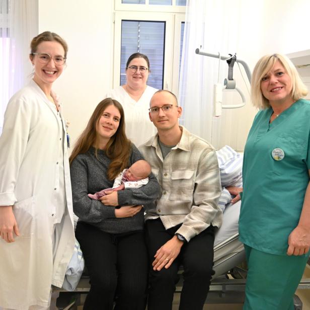 Eine Frau hält ein Neugeborenes im Arm, neben ihr sitzt ein Mann, umgeben von medizinischem Personal in einem Krankenhauszimmer.